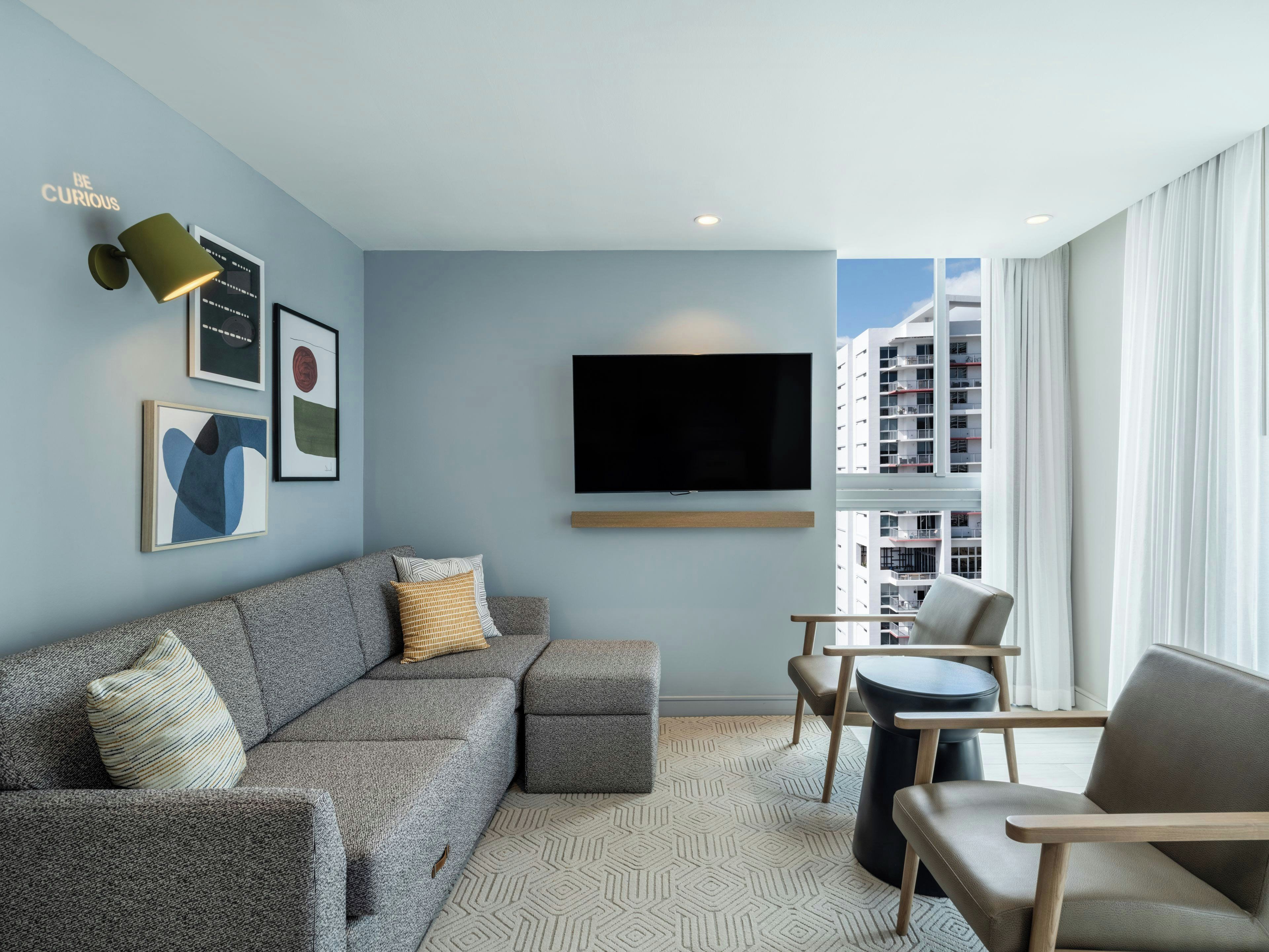 Una sala de estar moderna con un sofá seccional gris, un televisor de pantalla plana y vistas a la ciudad a través de grandes ventanales.
