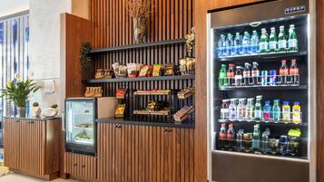 A vending machine stocked with beverages and snacks, a refrigerated display case, and a wooden panel wall with a potted plant.