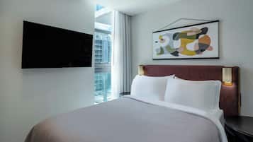 Habitación de hotel con una cama, un televisor de pantalla plana montado en la pared, una ventana con vistas a edificios y una obra de arte abstracta enmarcada.