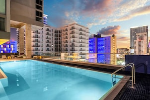 Una piscina en la azotea con barandilla de cristal, rodeada de edificios altos con balcones.