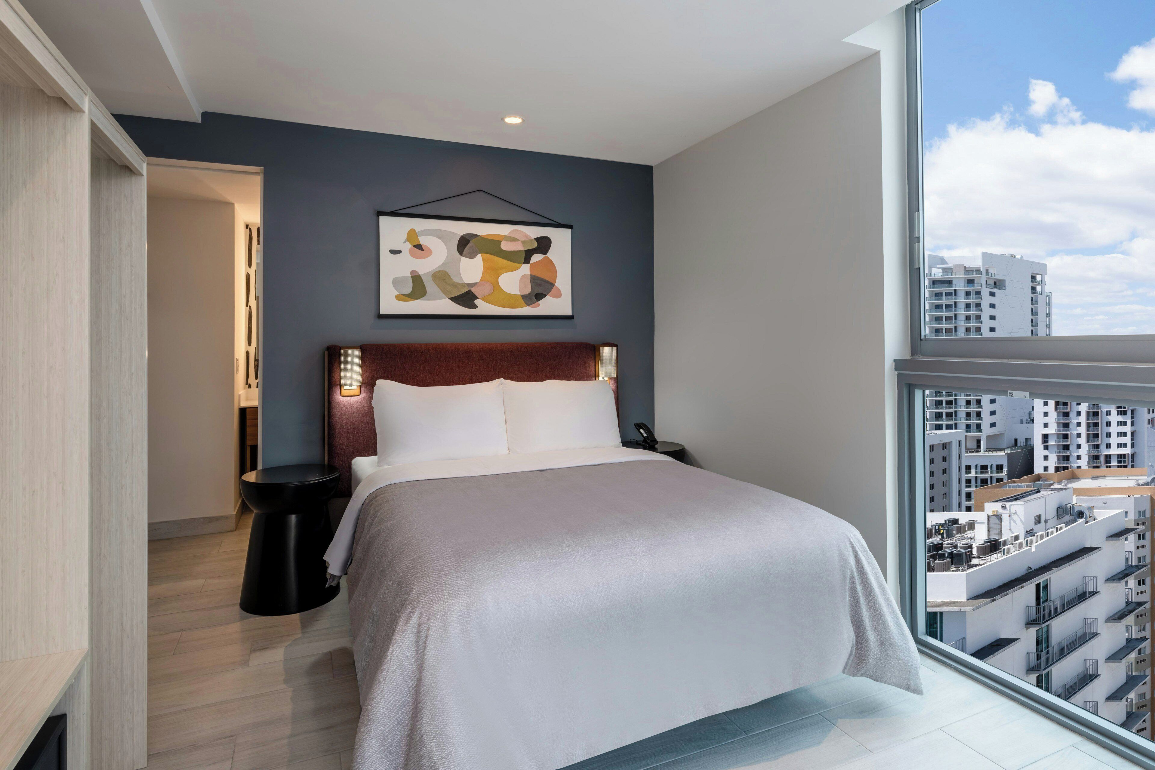 A modern hotel room with a large bed, a view of the city, and a contemporary abstract painting on the wall.