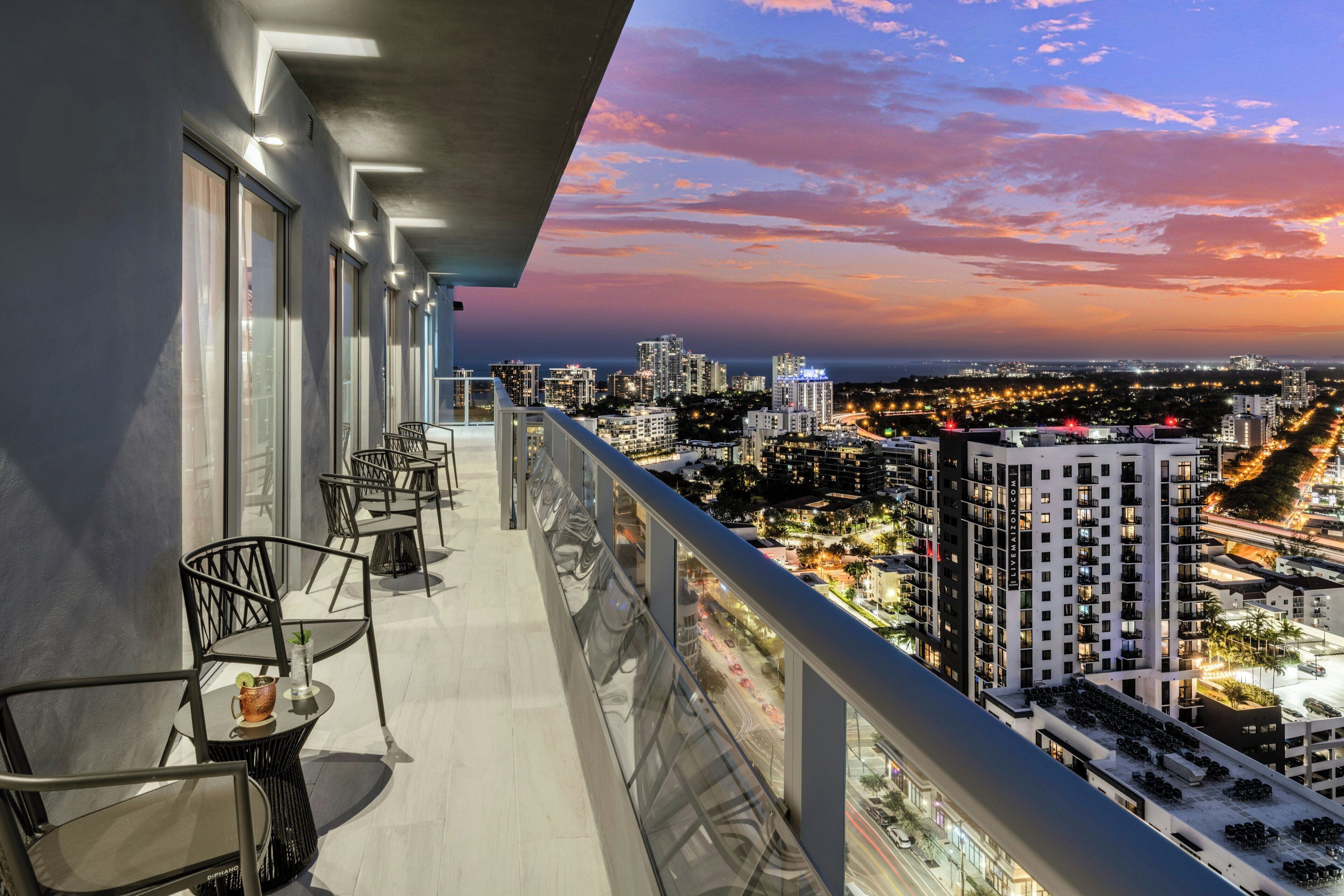 A balcony with outdoor seating, overlooking a cityscape at sunset.