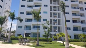 A multi-story white building with balconies, surrounded by palm trees and a well-maintained lawn. - espectacular apartamento (Playa Juan Dolio)