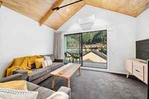 Een moderne woonkamer met een grijze bank, een houten salontafel en een tv-meubel. Een groot schuifglas leidt naar een terras met een prachtig uitzicht op een bebost gebied.