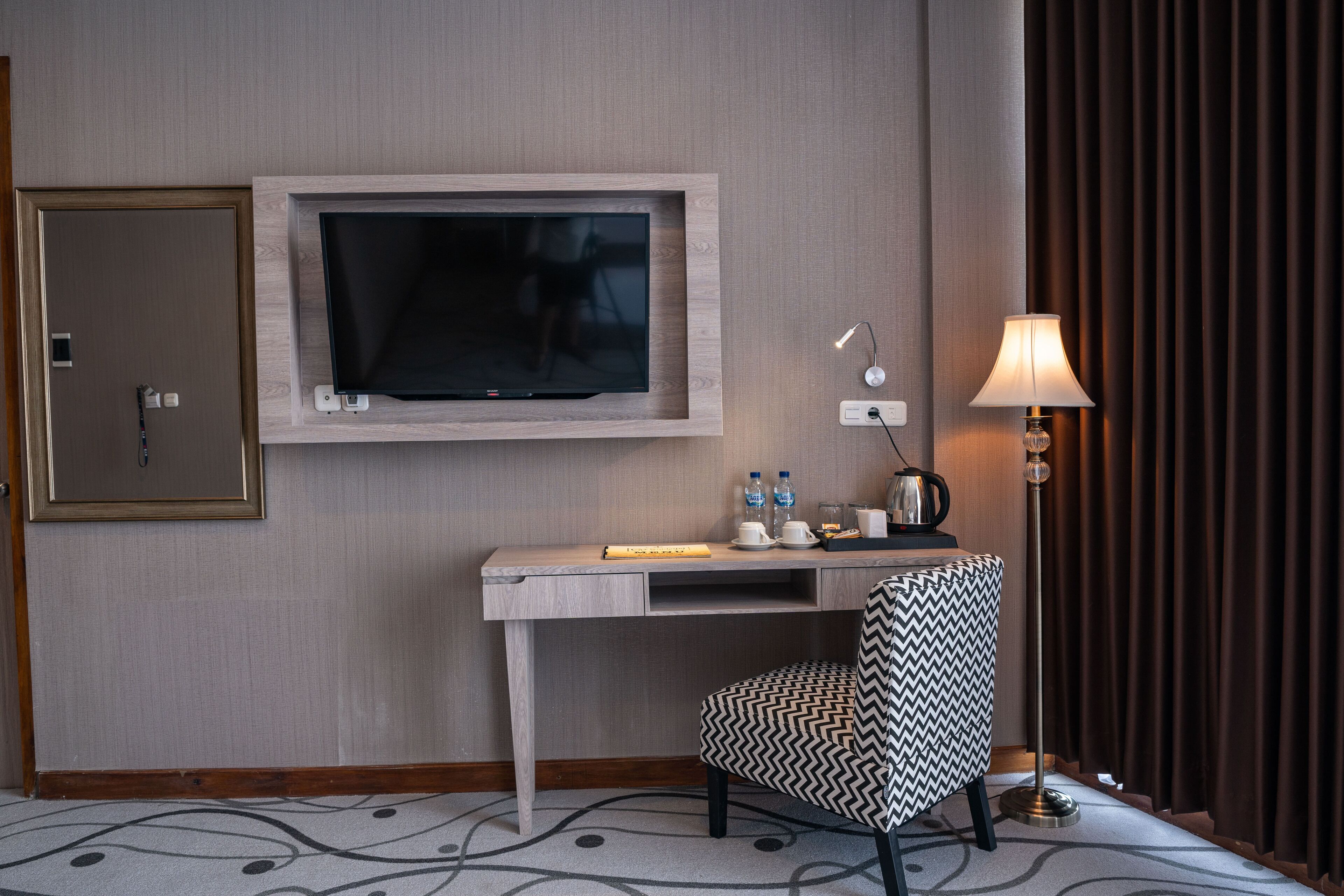 A hotel room with a flat-screen TV mounted on the wall, a desk with a chair, a lamp, and a mirror.