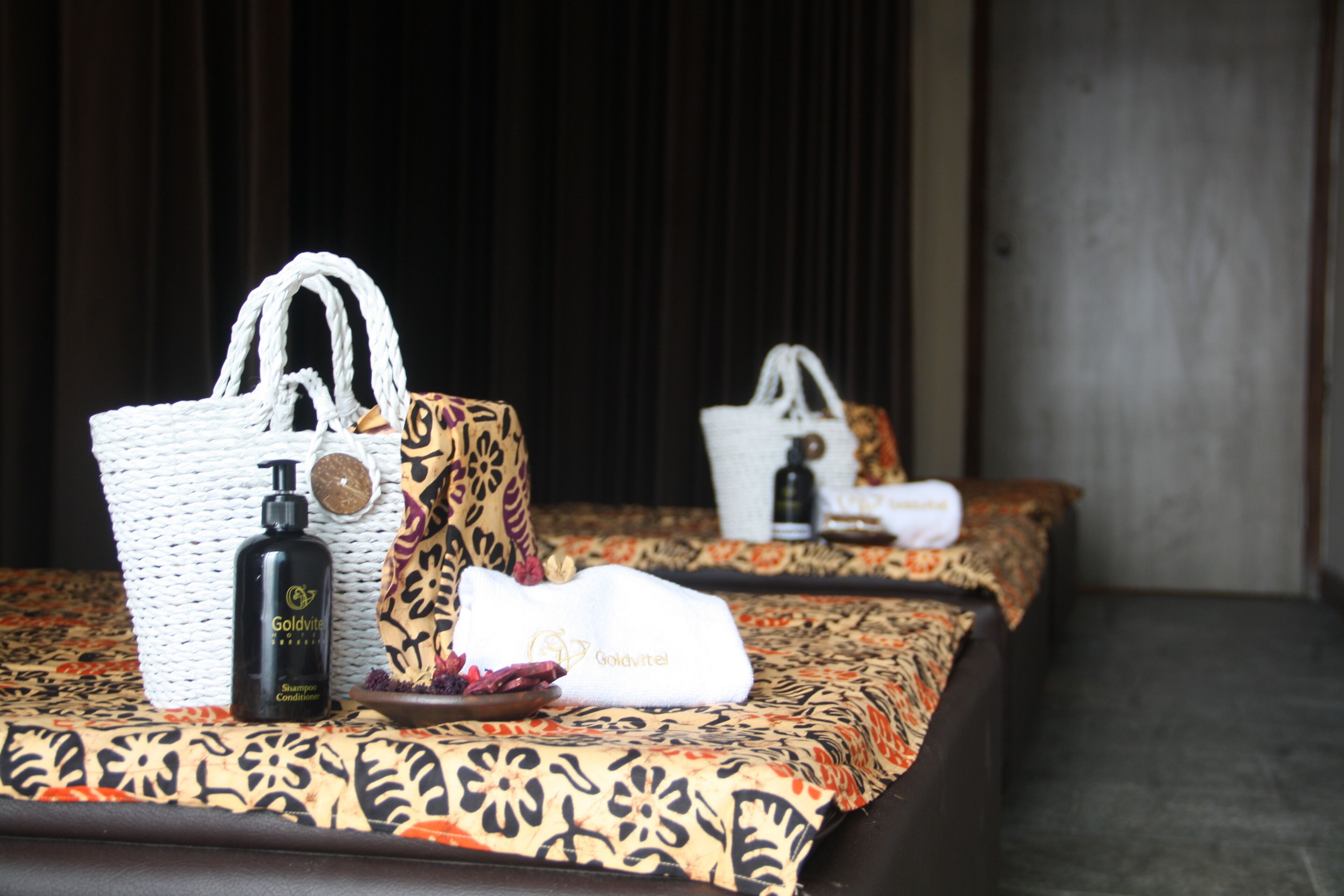 A hotel room with two beds, each with patterned bedding and a white woven bag on the bed. There is a bottle of Goldvital shampoo and conditioner on the first bed.
