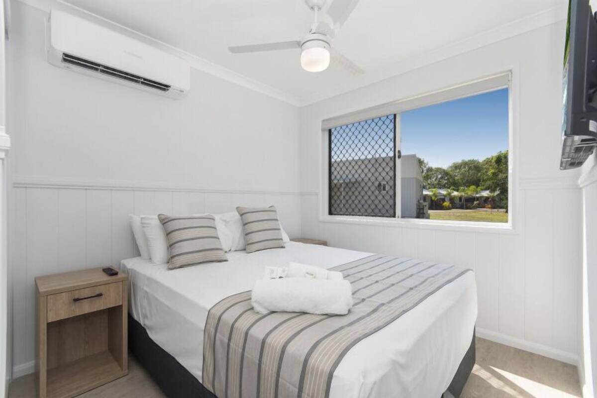 A bedroom with a bed, bedside table, air conditioning unit, ceiling fan, window with a view, and a TV mounted on the wall.