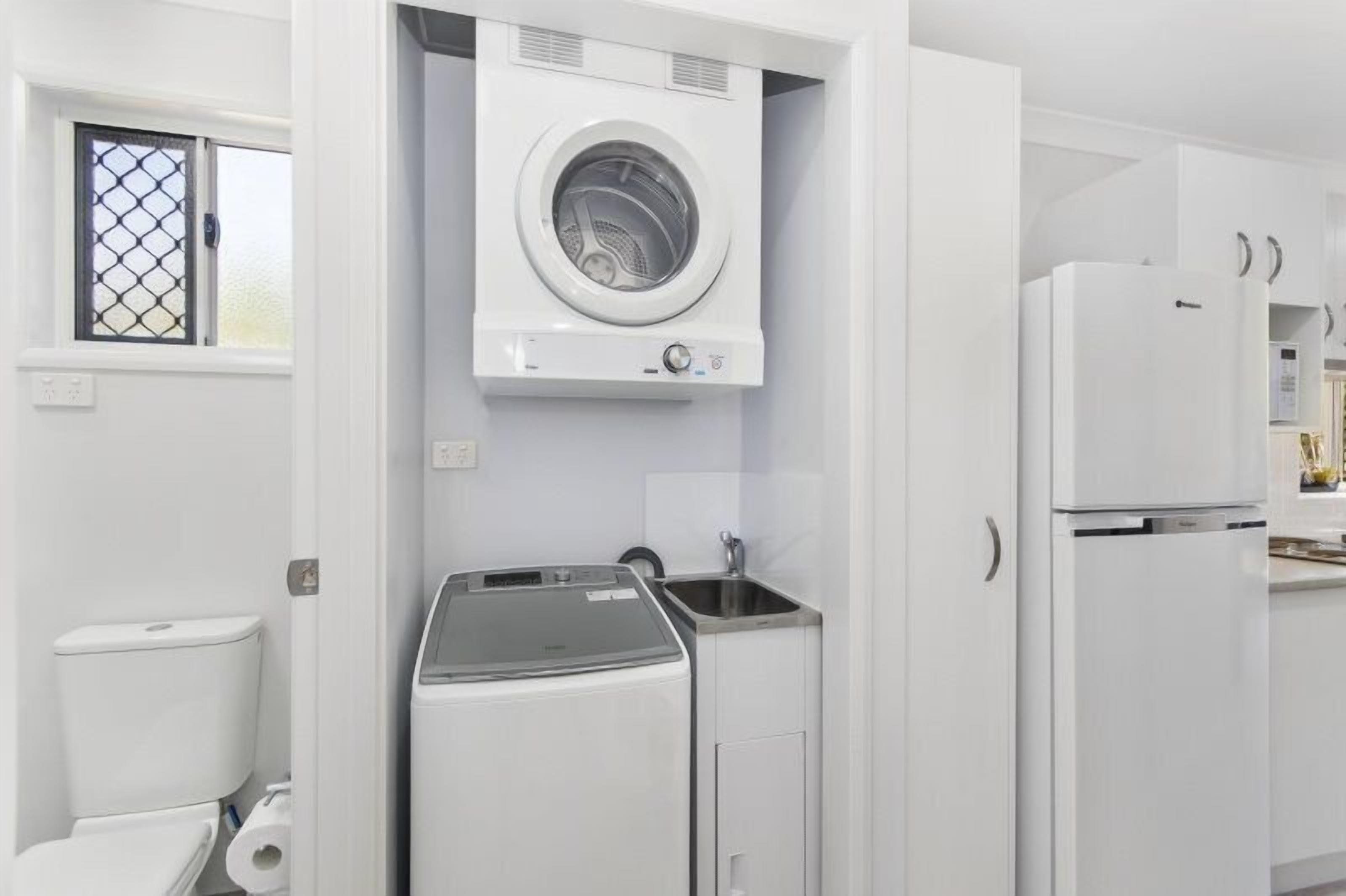 A compact laundry room with a front-loading washer and dryer, a sink, and a window with a metal screen.