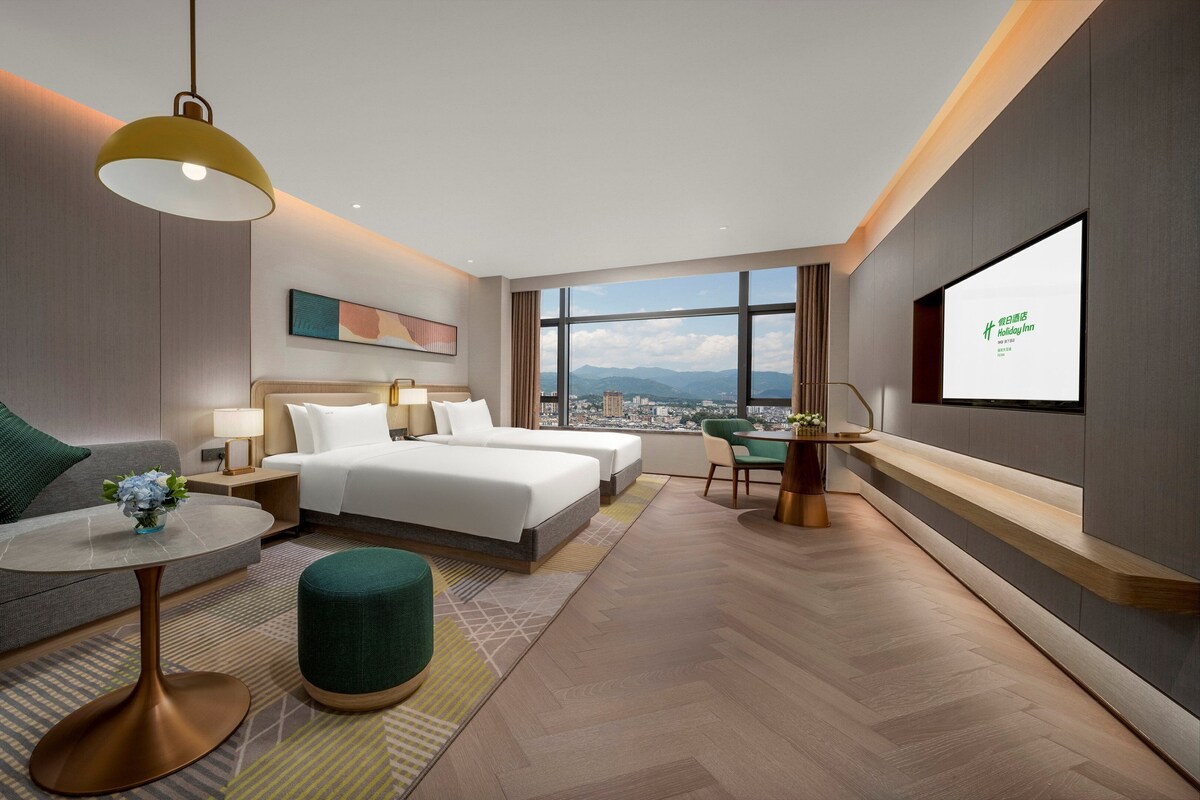 A modern hotel room with a city view, two beds, a sofa, a dining table, and a TV screen.
