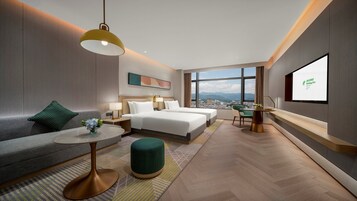 Habitación de hotel moderna con vistas a la ciudad, dos camas, un sofá, una mesa de comedor y una pantalla de televisión.