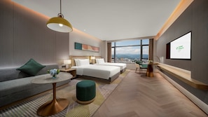 A modern hotel room with a city view, two beds, a sofa, a dining table, and a TV screen.
