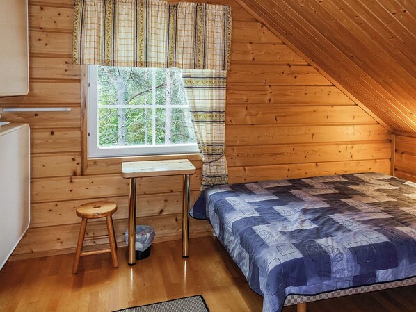 A small, cozy room with a wooden sloped ceiling, a single bed with a patterned blanket, a small table with a chair, and a window with curtains.