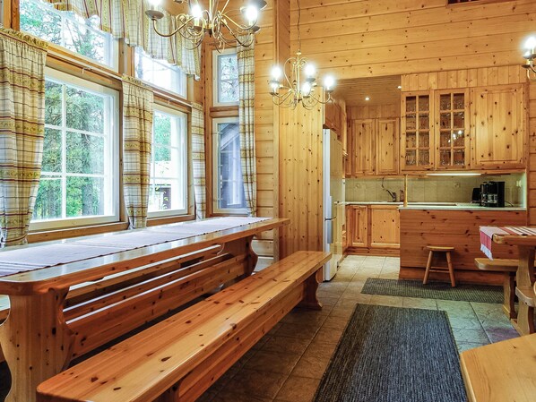 Interior de una cabaña de madera con mesa de comedor, bancos, zona de cocina y una araña.