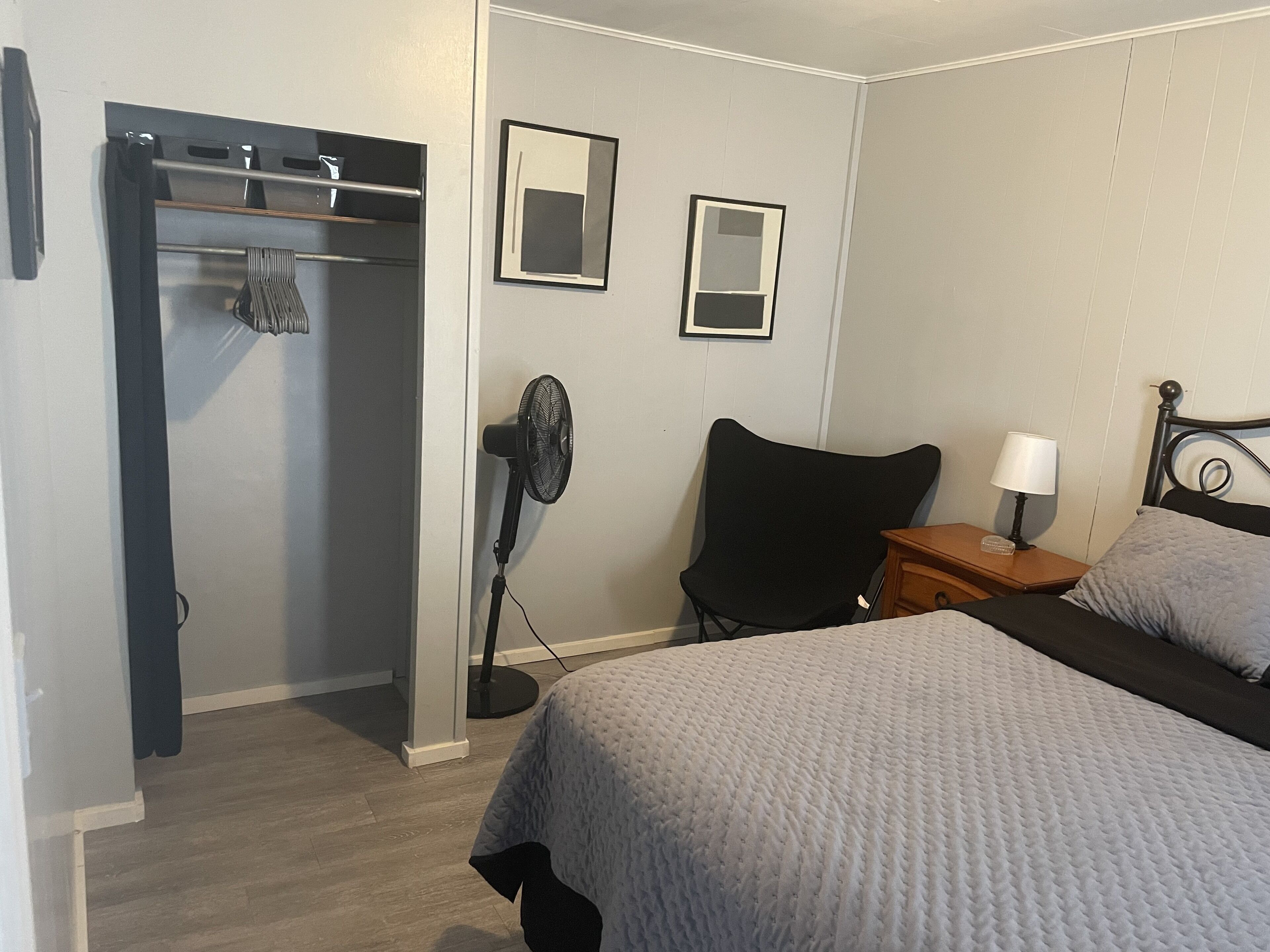 Une chambre à coucher avec un lit, une chaise, une table de chevet avec une lampe, un ventilateur et un porte-manteau avec des vêtements.