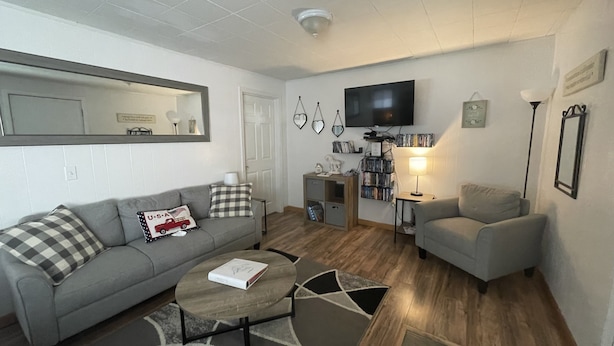 Una sala de estar con un sofá gris, una pequeña mesa redonda, un sillón gris, un televisor, una estantería y una lámpara.