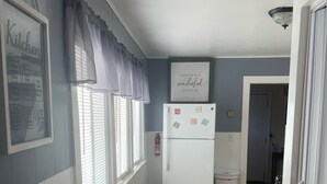 Una cocina con un refrigerador blanco, una ventana con persianas, un extintor de incendios y una puerta que da a otra habitación.