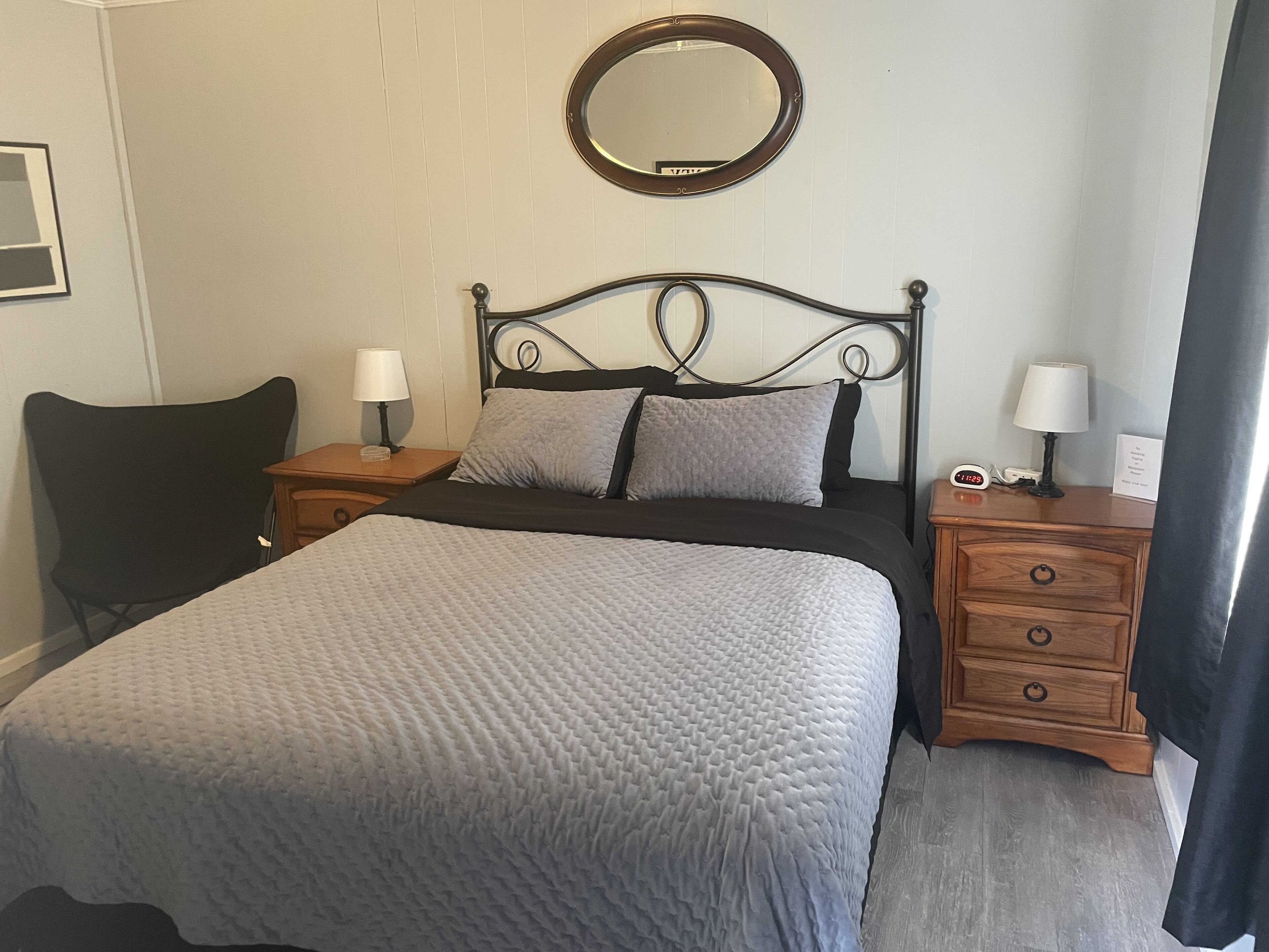 Un lit bien fait, avec une couverture grise, une tête de lit en métal noir et deux tables de chevet avec des lampes.