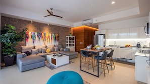Uma sala de estar moderna com um sofá cinzento, uma mesa de centro e um espaço de jantar com banquetas altas. Há uma cozinha com armários brancos e uma geladeira de aço inoxidável.