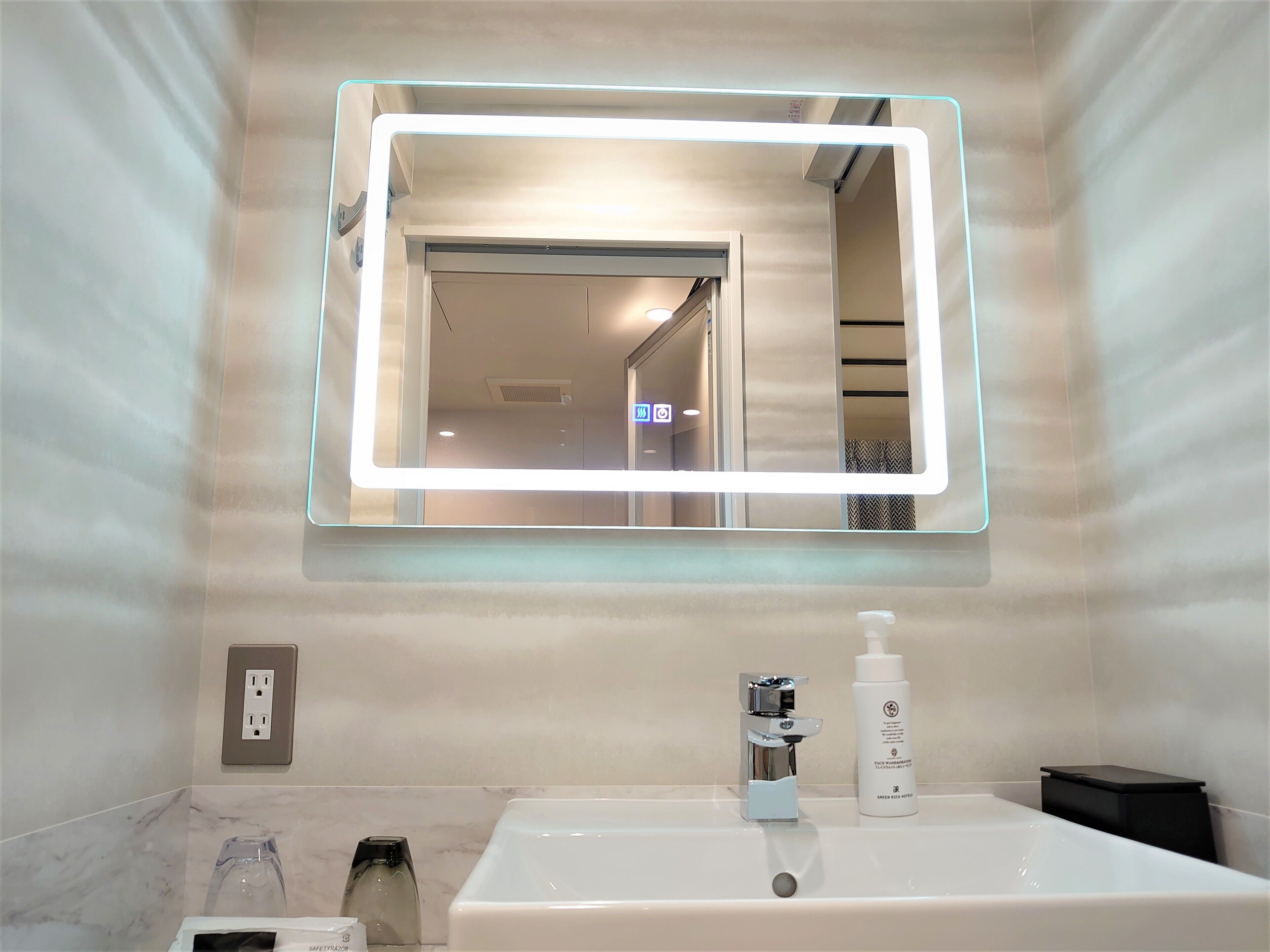 A bathroom with a large mirror, a sink, a bottle of shampoo, and a towel dispenser.