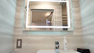 A bathroom with a large mirror, a sink, a bottle of shampoo, and a towel dispenser.