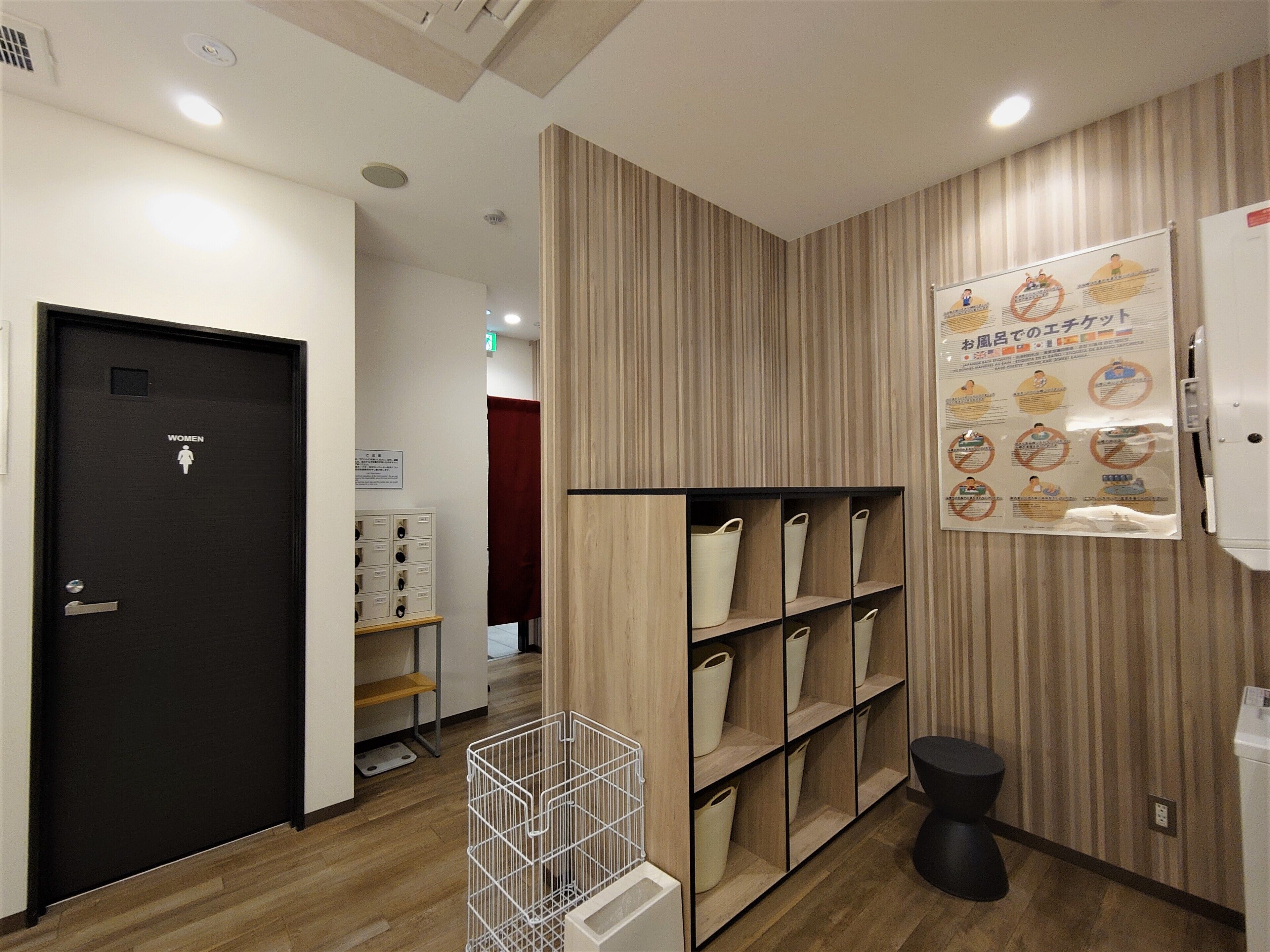 A hotel restroom with a wooden vanity, shelves with storage bins, a black toilet, and a wall poster with health and safety information.