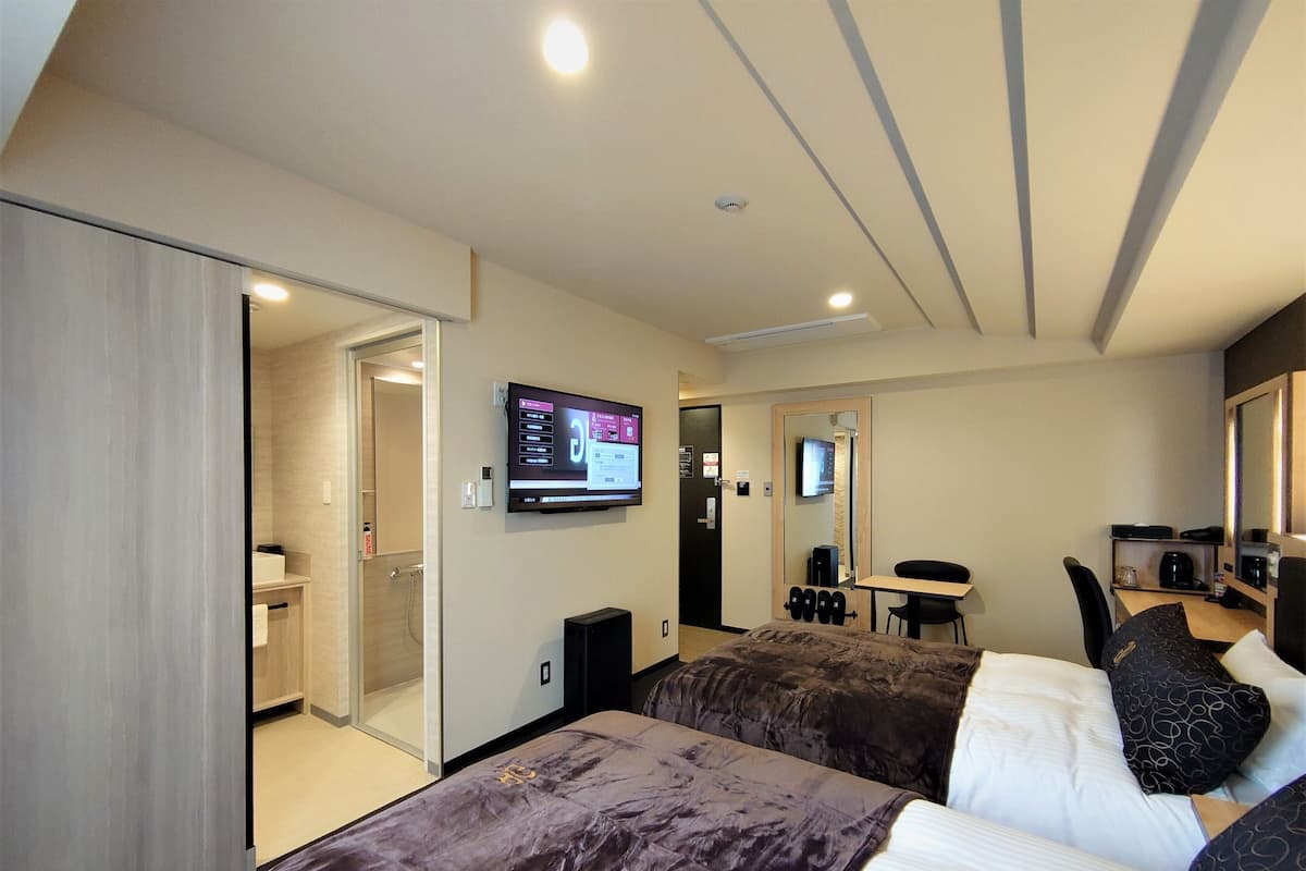 Hotelkamer met twee bedden, een aan de muur bevestigde televisie, een bureau met stoel, en een klein tafeltje met koffieapparaat.