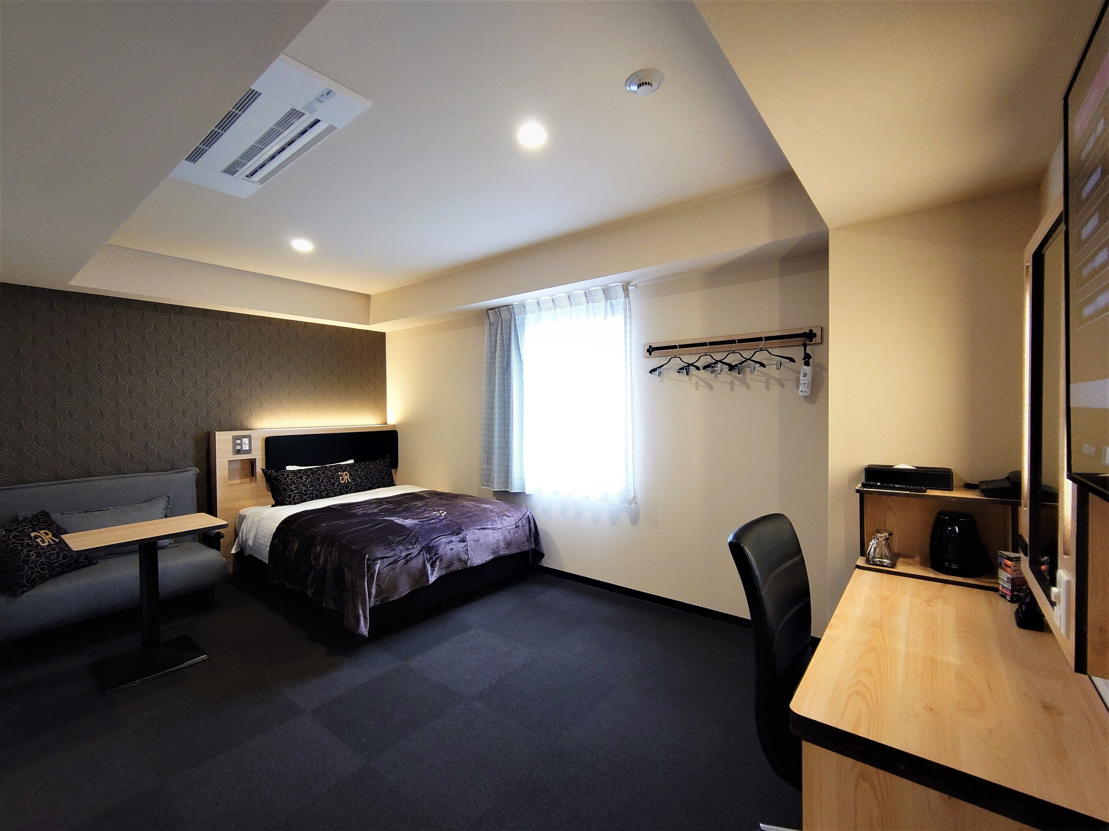 A hotel room with a bed, a desk, a chair, a coat rack, and a window with curtains.