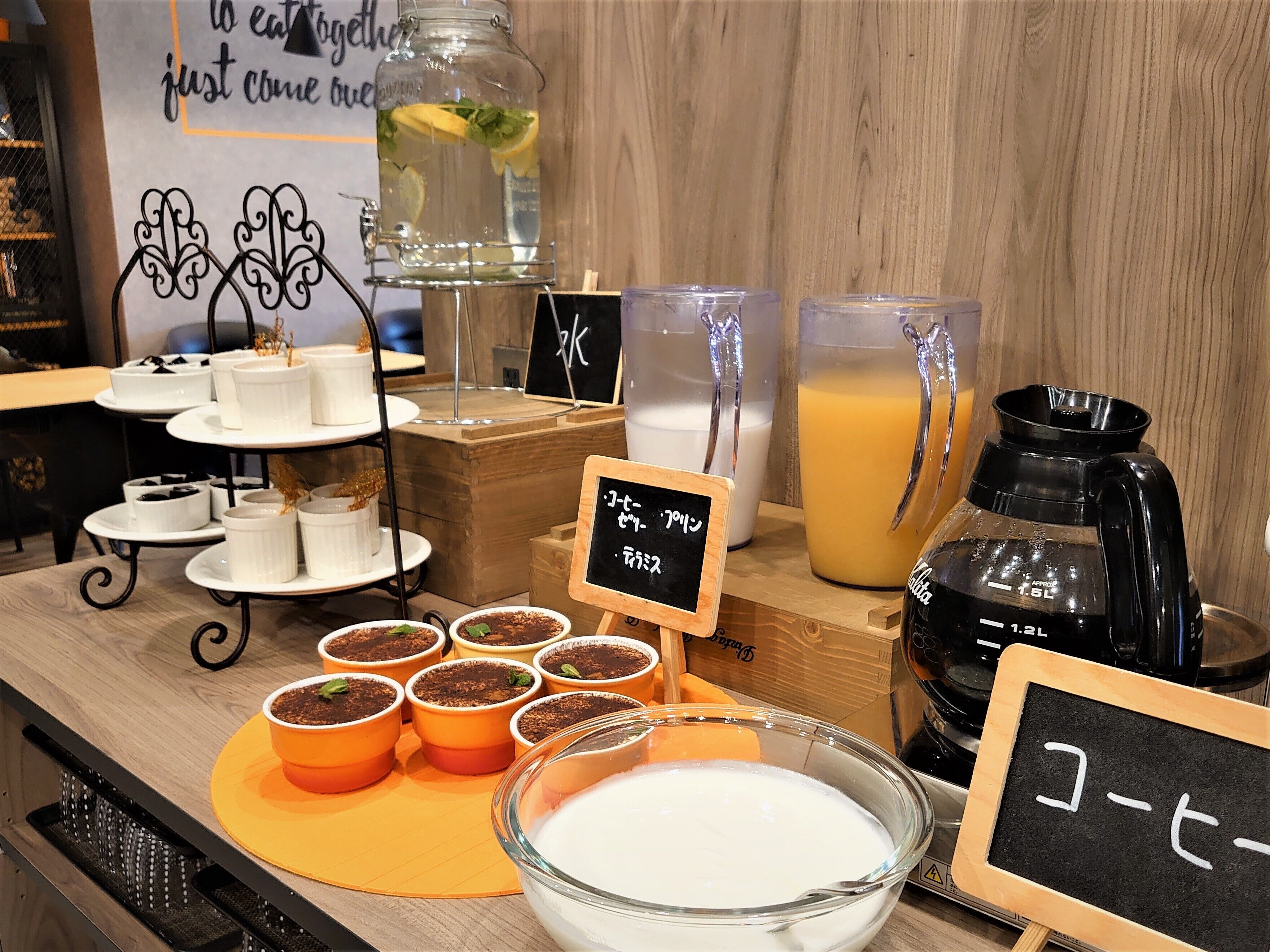 Una estación de café con diversas bebidas, incluyendo una jarra de jugo de naranja, un tarro de vidrio con una rodaja de limón y varias tazas pequeñas de café.