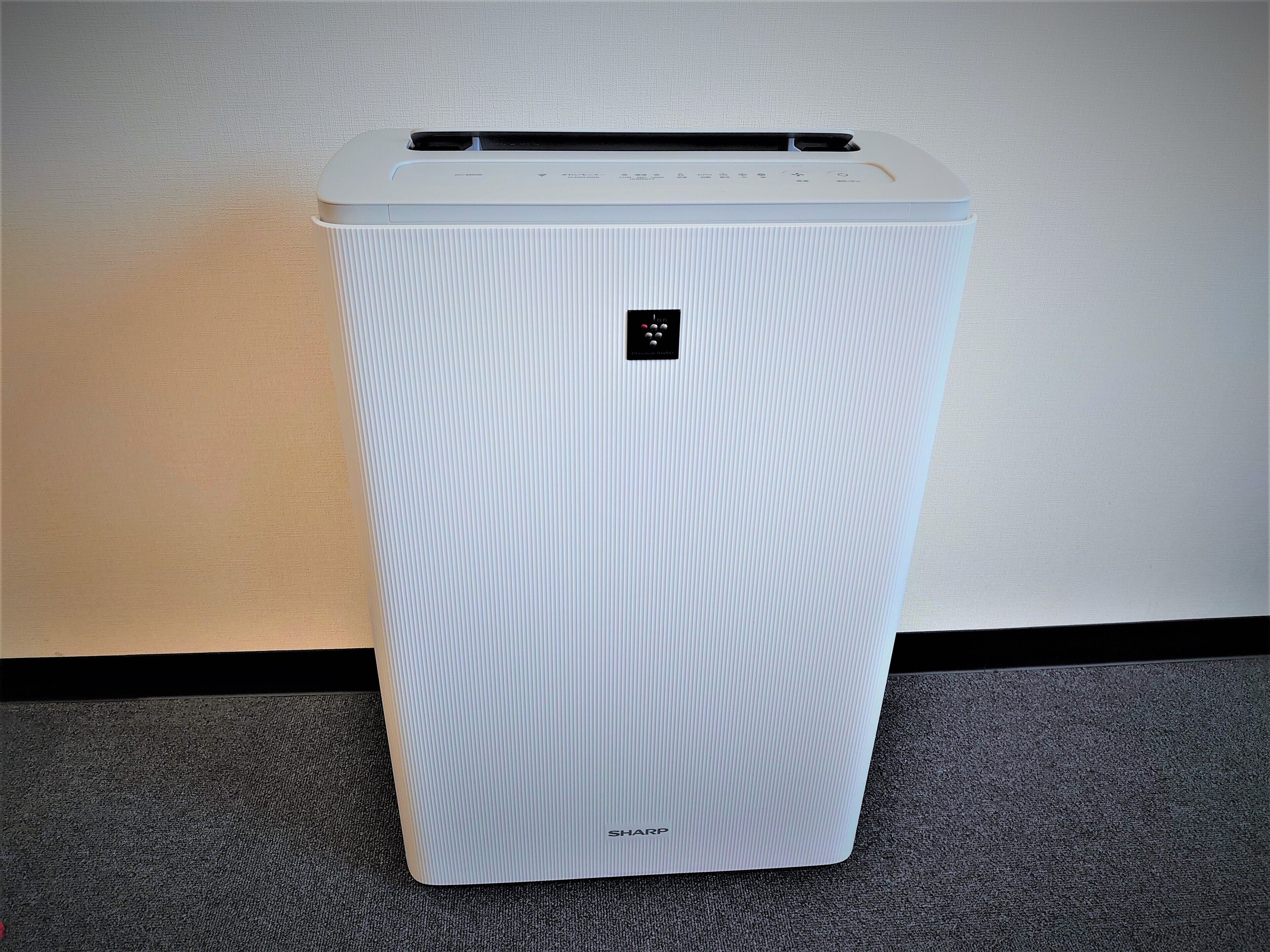 Un equipo de aire acondicionado blanco, con diseño acanalado y forma rectangular, con un panel de control en la parte superior y el nombre de la marca 'SHARP' en la parte inferior.