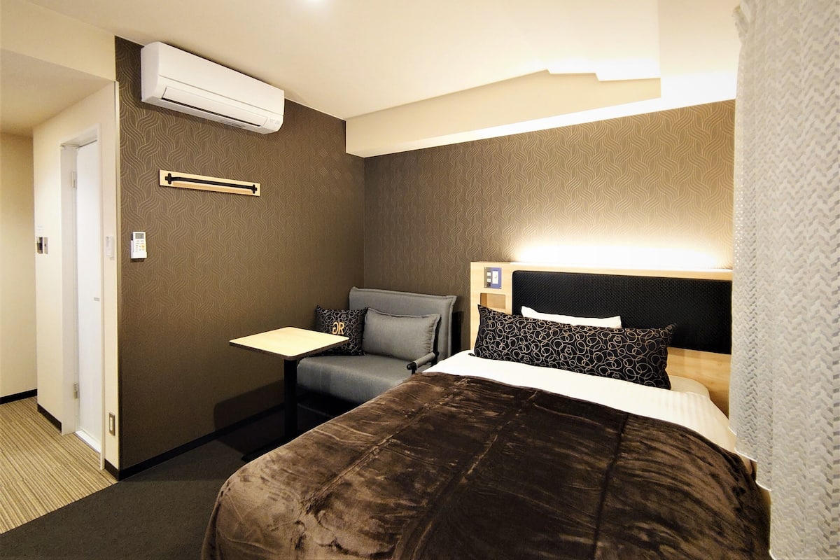 Een hotelkamer met een bed, nachtkastje en bureau. Er is een airconditioner aan de muur bevestigd.