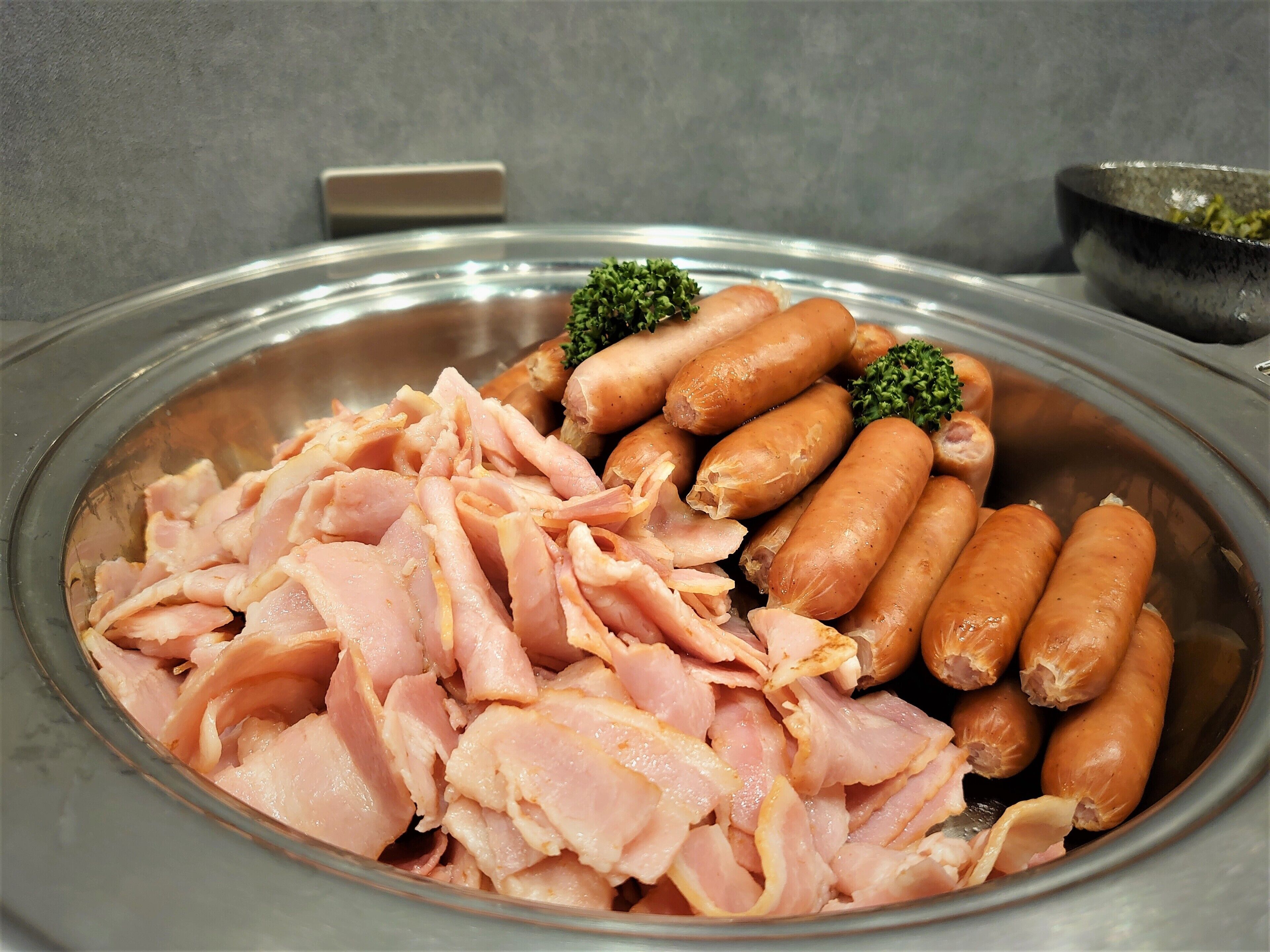 A stainless steel serving dish with sliced ham and sausages, garnished with broccoli.