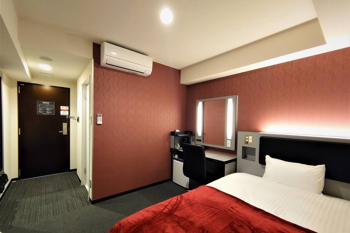 Een hotelkamer met een bed, bureau, stoel, spiegel, airconditioning en een deur met een bordje.