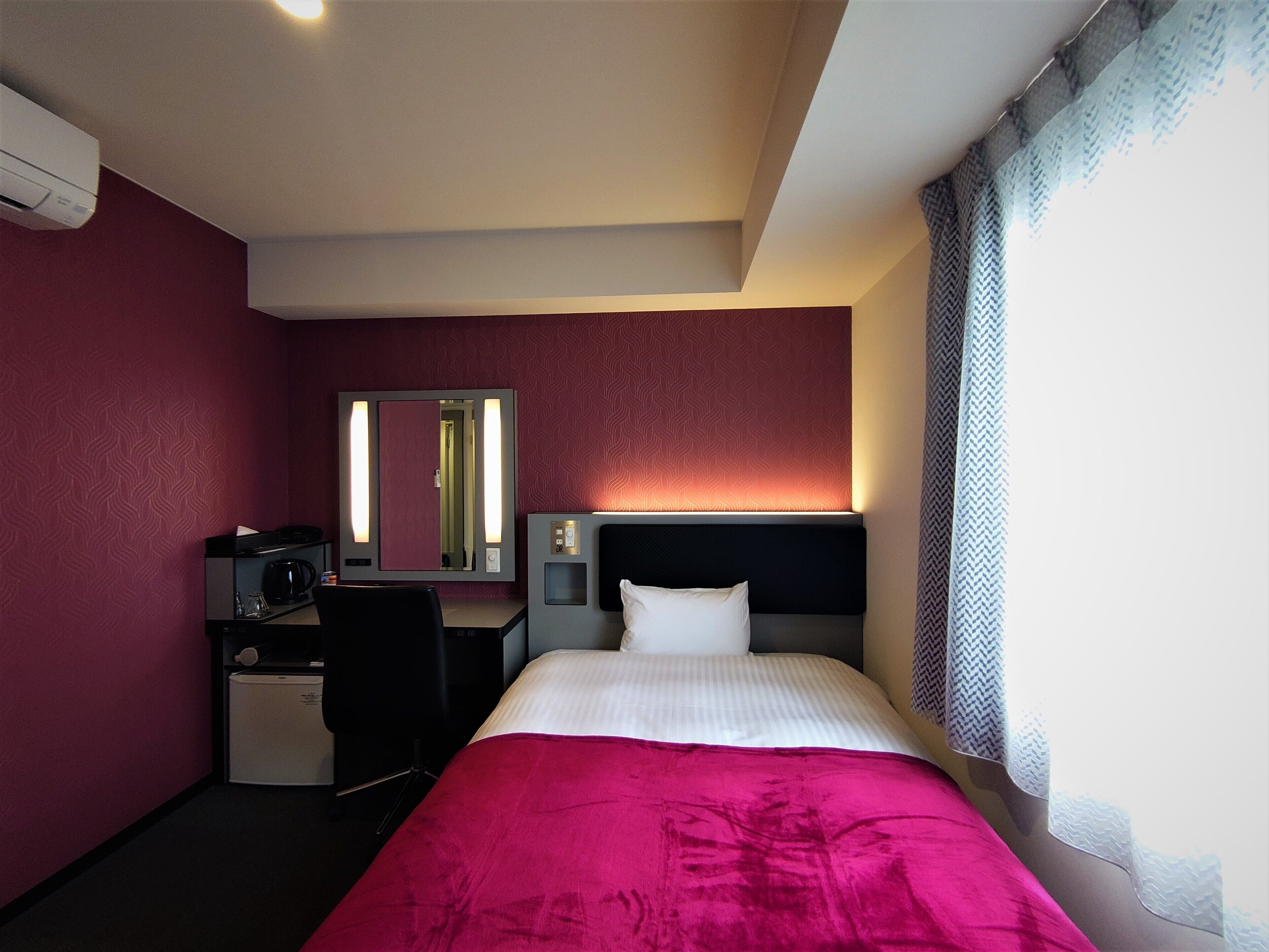 Habitación de hotel con cama, escritorio, silla, mesita, espejo y ventana con cortinas.