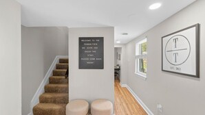 A hallway with a staircase, two ottomans, a welcome sign, and a framed logo on the wall. - * Beautiful Oasis w/ No Detail Spared * (Lutherville-Timonium)