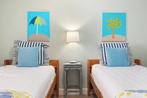 Une chambre avec deux lits, chacun agrémenté d’oreillers à rayures et d’une couvre-lit à motifs bleus et blancs. Une petite table avec une lampe se trouve entre les deux lits.