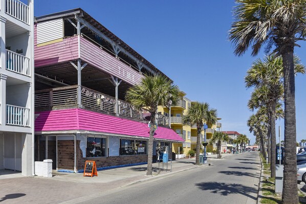 Un immeuble à plusieurs étages, doté d’une marquise rose et de balcons, situé dans une rue ensoleillée bordée de palmiers.