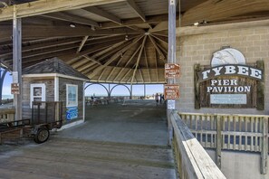 Ein Pier-Pavillon mit Holzdach, Schilder, die Fahrräder, Tiere, Motorradfahrer und Skater verbieten, und ein Schild, das angibt, dass es Teil des Tybee Island Pier Pavilion ist.
