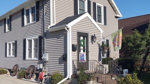 A two-story house with a gabled roof, a front porch, and a small garden area with a wreath and a flag. - Tina's bungalow  (Manitowoc)