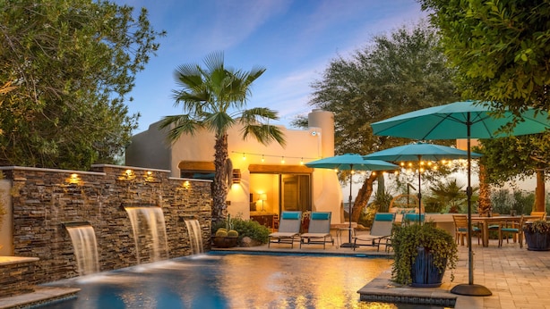 Zona de piscina con cascada, asientos exteriores y un edificio con iluminación cálida.