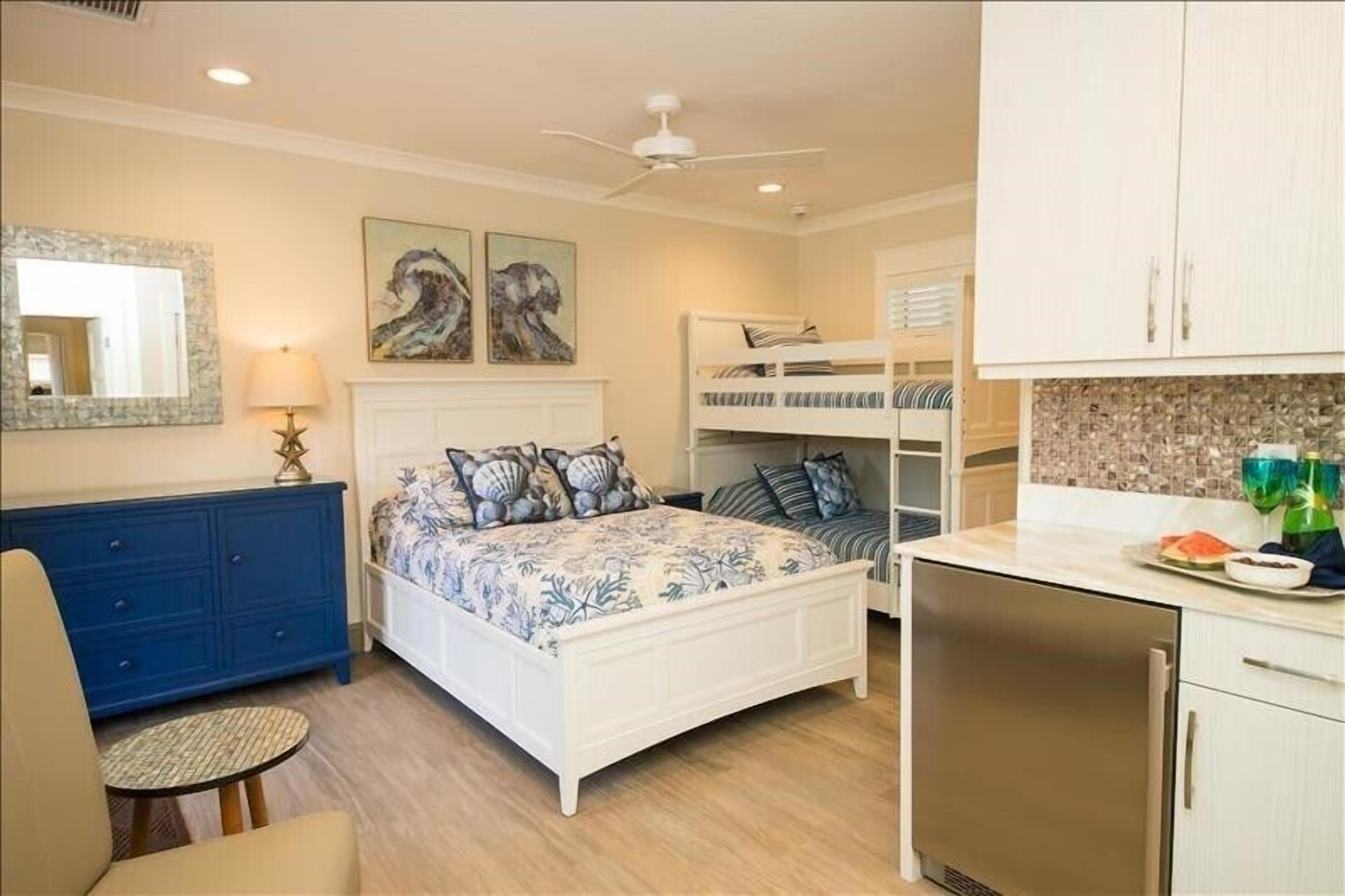 Une chambre à coucher comprenant un lit, un bureau, une chaise, une commode, un ventilateur de plafond et un coin cuisine.