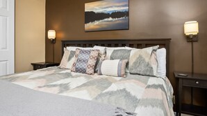 Quarto com cama, mesa de cabeceira, luminária na parede e pintura de uma paisagem.