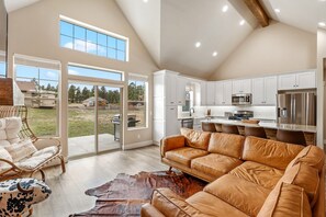 A spacious living room with a large leather sofa, a dining area with a long table, and a kitchen with white cabinets and stainless steel appliances. - Rustic Home - Central AC, Game room, 4BR, Sleeps10 (Williams)