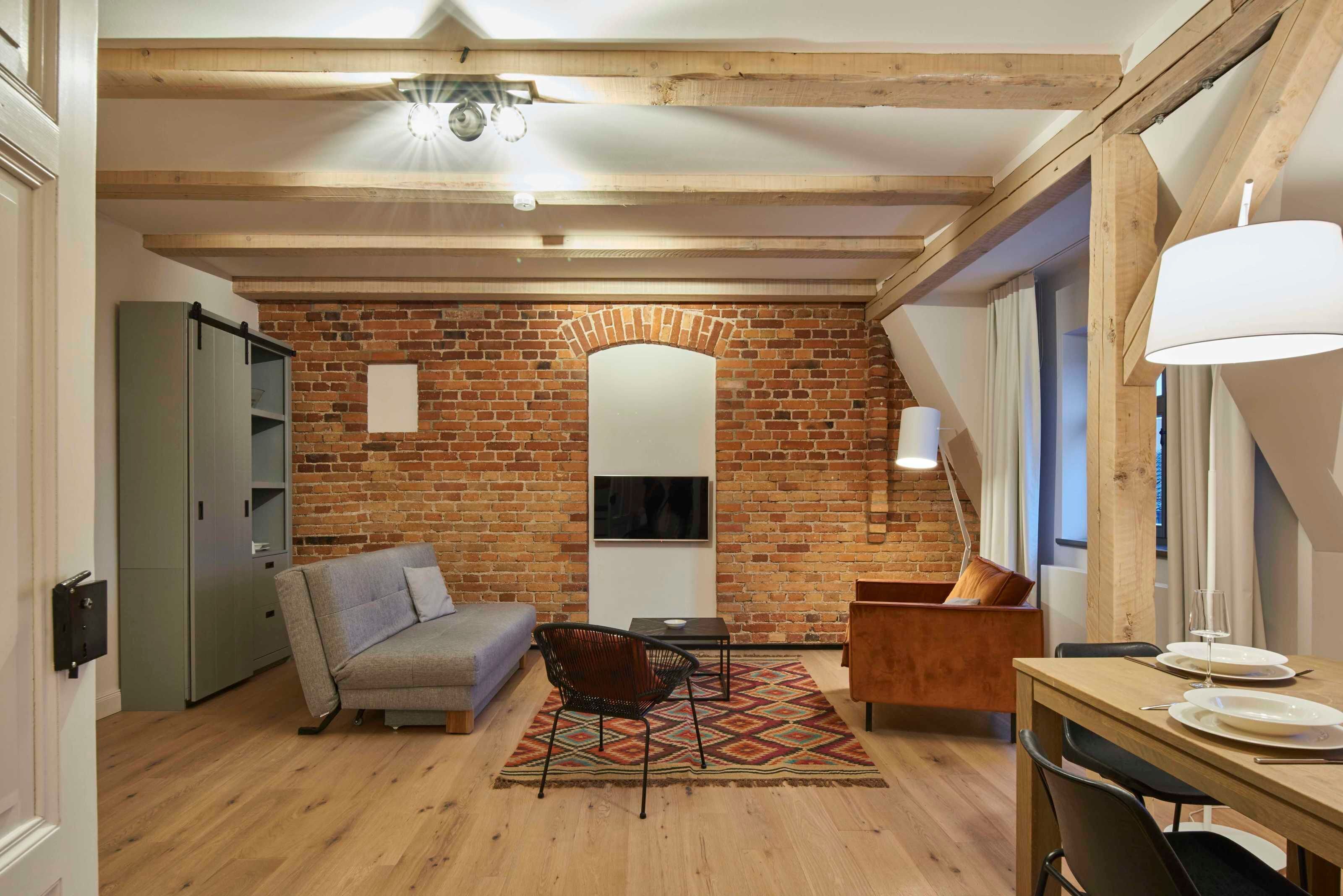 Ein gemütliches Wohnzimmer mit Backsteinwand, Holzbalken, einer Couch, einem Sessel, einem kleinen Tisch und einer Essecke mit Lampe und Tellern.