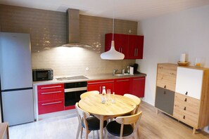 A compact kitchen with a microwave, oven, and red cabinets. There is a wooden dining table with chairs and a hanging light fixture. - Apartment Remise - Villa Inga & Anne (Heringsdorf)