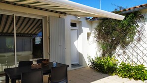 Un patio con una mesa y sillas, una puerta corrediza de vidrio, una pared blanca con un cerramiento de celosía y follaje.