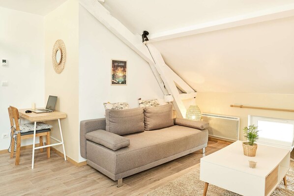 Une chambre à coucher confortable, aménagée en mezzanine, avec un plafond en pente, un bureau en bois avec un ordinateur portable, un canapé gris et une table de chevet blanche agrémentée d’une plante.