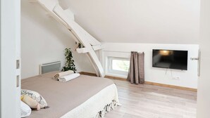 A bedroom with a sloped ceiling, a bed with a white and beige bedspread, a potted plant, a window with curtains, and a flat-screen TV mounted on the wall.