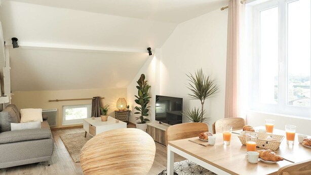 A modern living room with a sofa, a round wooden ottoman, a dining table with breakfast items, a TV, and a large window with curtains.