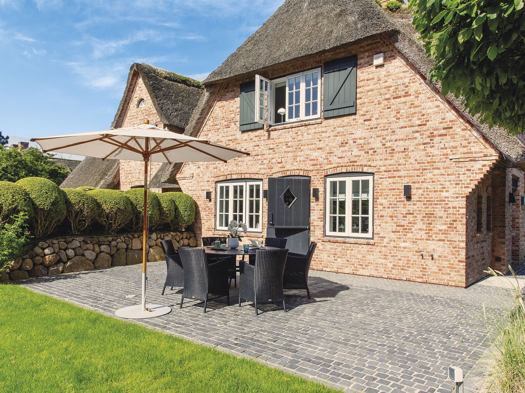 A brick house with a thatched roof, a patio with outdoor furniture, and a stone wall.