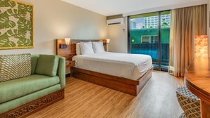 A hotel room with a bed, a green sofa, a wooden coffee table, and a large window with a view of buildings. - Climate Controlled Studio - Bamboo Waikīkī Hotel #409 (Honolulu)